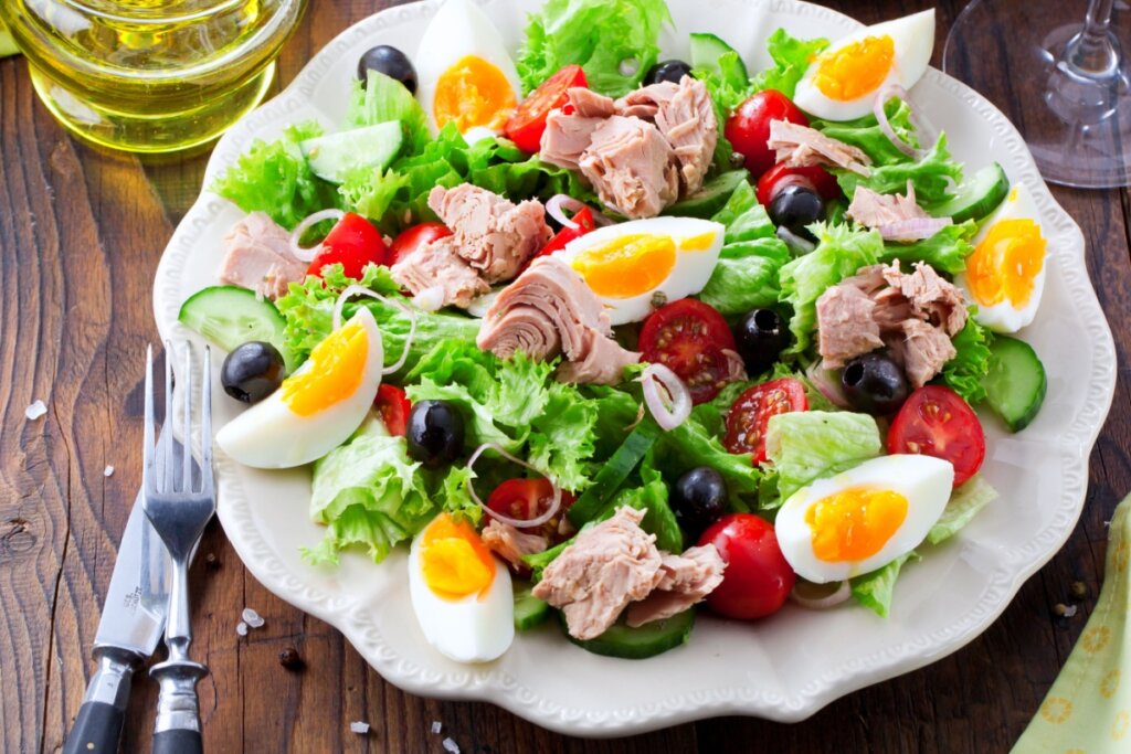 Salada de atum com ovos e legumes servido em prato branco em cima de mesa de madeira