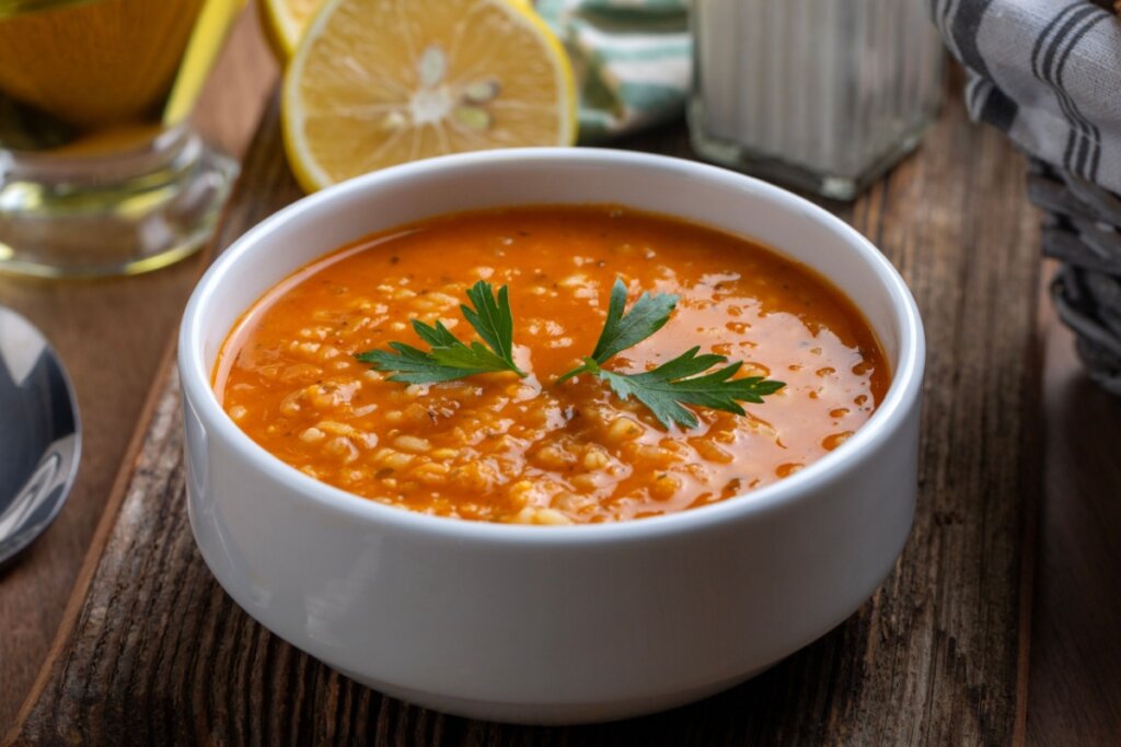 A imagem mostra uma tigela branca com uma sopa de cor alaranjada, com textura cremosa e levemente granulada, sugerindo a presença de grãos ou ingredientes triturados. No centro, há folhas de salsinha usadas para finalizar e decorar o prato. A tigela está apoiada sobre uma superfície de madeira rústica, que traz um aspecto acolhedor à cena.