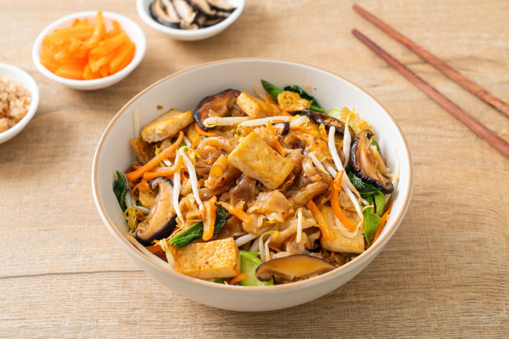 Tofu, espinafre, macarrão, cogumelos, repolho, acelga e cenoura em um recipiente branco em mesa de madeira