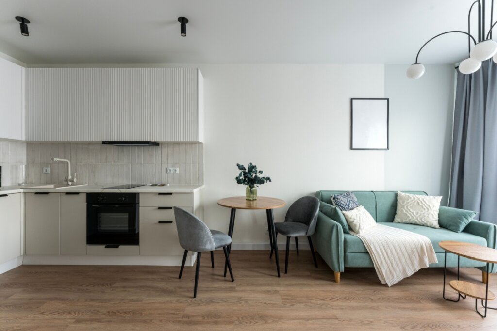 Visão frontal de um apartamento integrado em estilo estúdio contemporâneo e minimalista. À esquerda, há uma cozinha compacta com armários planejados em tom off-white e detalhes ripados na parte superior, equipada com cooktop, forno embutido preto e uma torneira branca sobre a bancada clara. No centro, uma pequena mesa redonda de madeira com pés palito pretos é acompanhada por duas cadeiras estofadas em veludo cinza. À direita, um sofá de três lugares em tom verde-menta decorado com almofadas texturizadas e uma manta clara. Um quadro com moldura preta e tela em branco decora a parede branca. O chão é revestido de piso vinílico amadeirado claro.