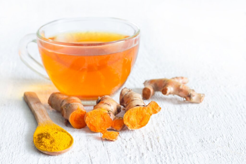 Chá de cúrcuma servido em xícara de vidro com pedaços de cúrcuma ao lado e uma colher de pau com cúrcuma em pó