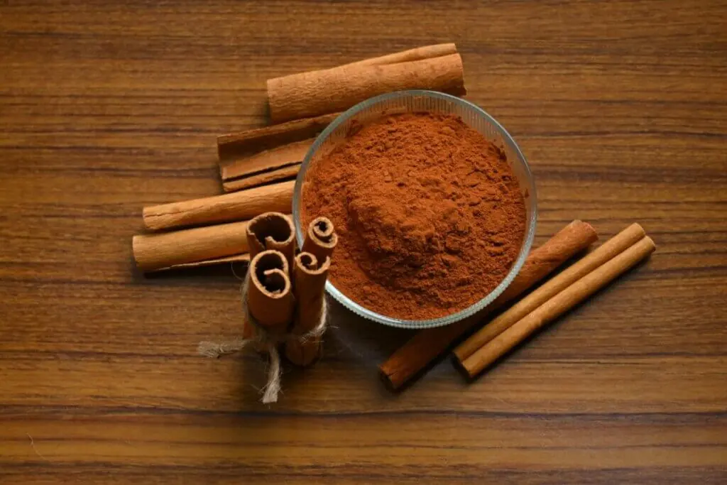 Uma composição sobre uma superfície de madeira marrom mostrando canela em duas formas: em pó e em pau. No centro, há uma pequena tigela de vidro redonda cheia de canela em pó de cor marrom-avermelhada. Ao redor da tigela, estão espalhados vários paus de canela secos. No canto inferior esquerdo, três paus de canela estão amarrados verticalmente com um pedaço de barbante rústico.