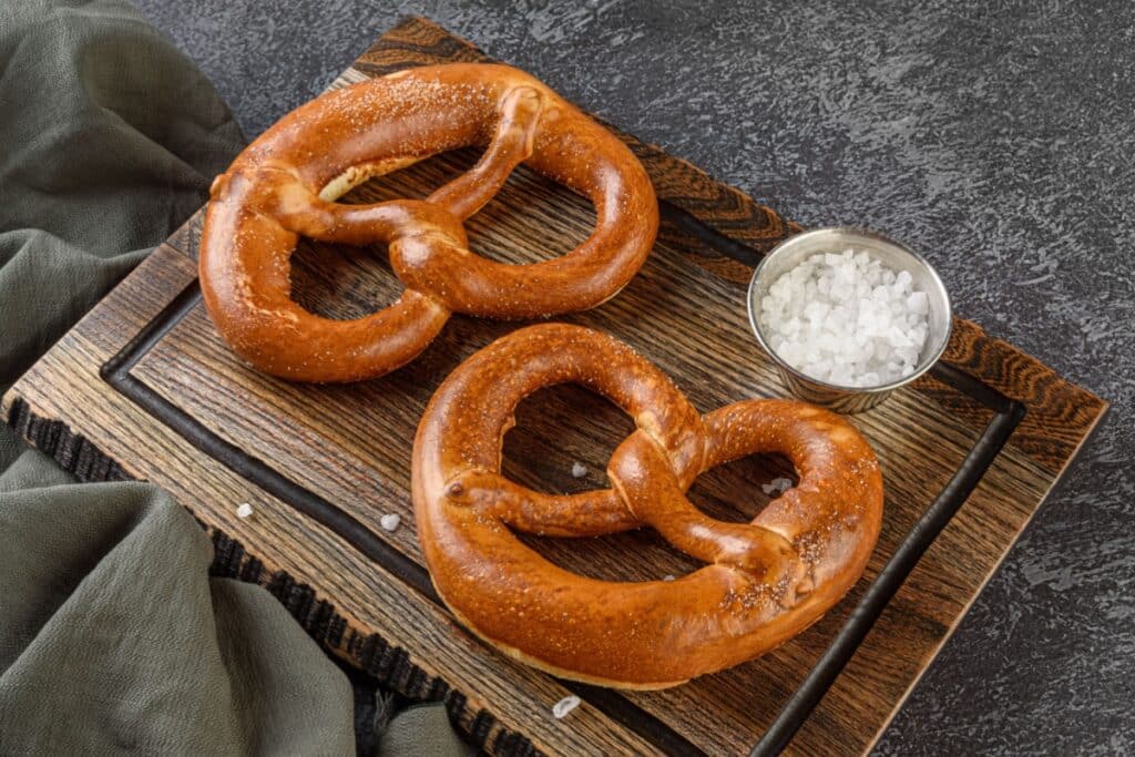 Dois pretzels grandes, de massa dourada e brilhante no formato tradicional de nó entrelaçado, estão dispostos sobre uma tábua retangular de madeira escura com texturas de veios evidentes. No canto superior direito da tábua, há um pequeno ramequin de metal cheio de cristais de sal grosso. A tábua repousa sobre uma superfície texturizada cinza-escura, acompanhada por um pano de tecido verde-oliva amassado no canto inferior esquerdo.