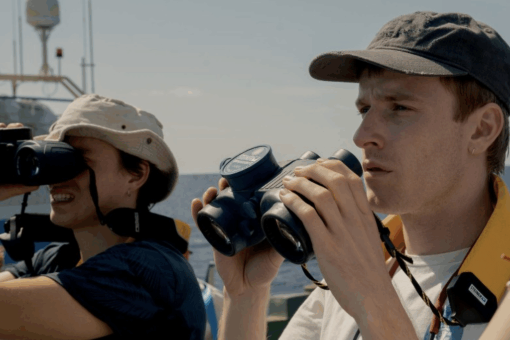 Dois jovens voluntários a bordo de um barco de resgate em alto-mar olham atentamente através de binóculos, procurando por refugiados no Mar Mediterrâneo sob a luz do dia.
