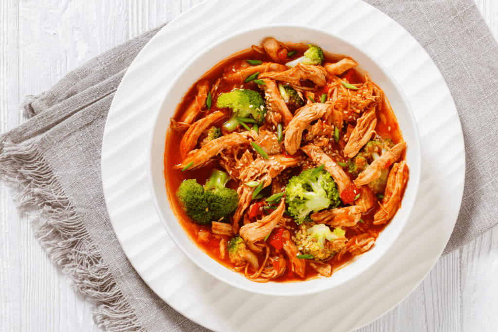 Vista superior de uma tigela branca com ensopado de frango desfiado ao molho vermelho, misturado com pedaços de brócolis e salpicado com gergelim e cebolinha. A tigela está sobre um prato branco maior e um pano cinza, dispostos em uma mesa de madeira clara