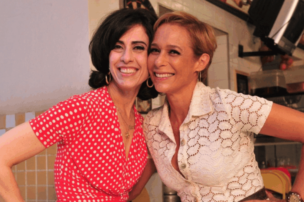 Uma foto de Tapas & Beijos apresentando as duas personagens principais, Fátima e Sueli, abraçadas e sorrindo calorosamente para a câmera. Fátima, à esquerda, tem cabelo preto curto e usa um vestido vermelho de bolinhas brancas e brinco de argola dourado. Sueli, à direita, tem cabelo loiro curto e usa uma camisa branca rendada abotoada e brinco de argola dourado. Elas estão em um ambiente de bar ou restaurante.