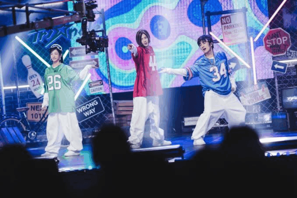 Três jovens dançam em um palco decorado no estilo urbano e hip-hop. Todos usam calças brancas largas e camisas de estilo esportivo numeradas: uma mulher à esquerda de verde (número 26), uma pessoa ao centro de vermelho (número 44) e um homem à direita de azul (número 34).
