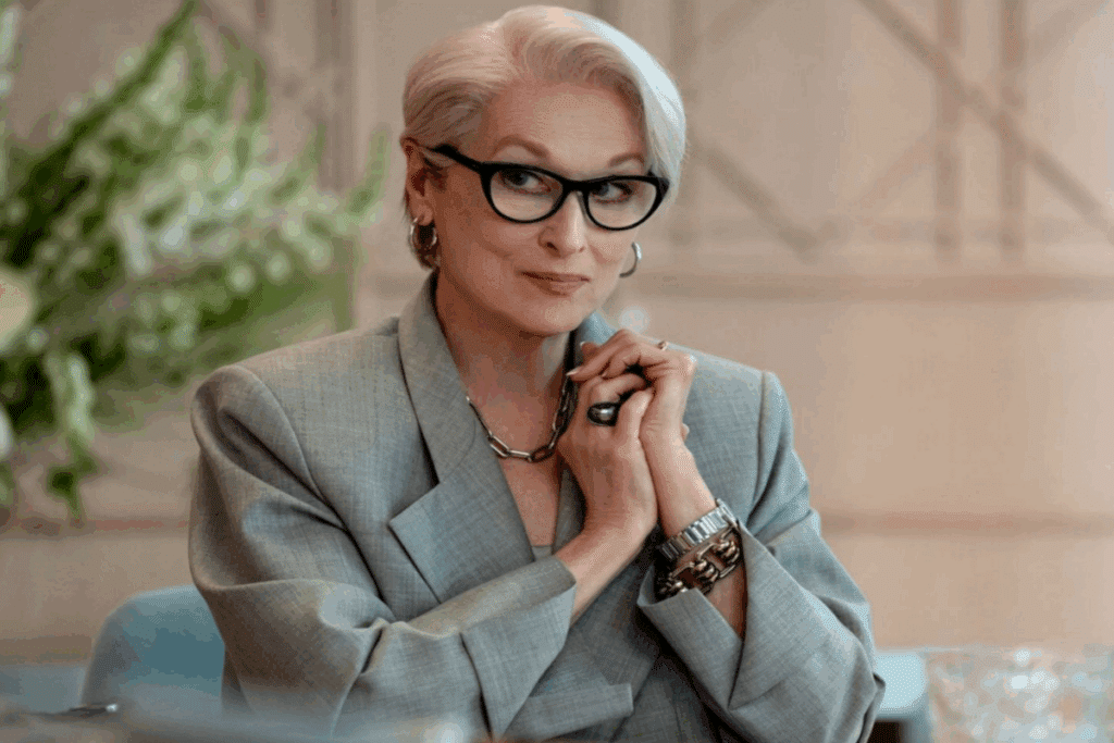Mulher com cabelo branco e curto, usando óculos preto e blazer cinza com mãos juntas perto do ombro