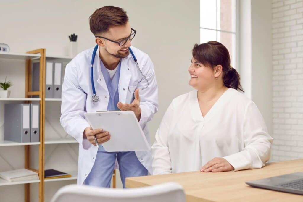 médico de roupa azul e jaleco branco, segurando prancheta, ao lado de mulher de blusa branca sentada e sorrindo
