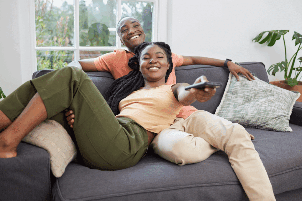 Um homem e uma mulher negros sorriem felizes em um sofá cinza. A mulher está deitada de forma descontraída, apontando o controle remoto em direção à tela, enquanto o homem está sentado logo atrás dela, apoiando o braço no encosto do sofá.