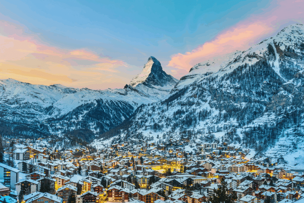Vista geral da cidade alpine de Zermatt ao anoitecer, com luzes acesas nas casas e chalés cobertos de neve, tendo a imponente montanha Matterhorn se destacando ao fundo sob um céu em tons de rosa e dourado.