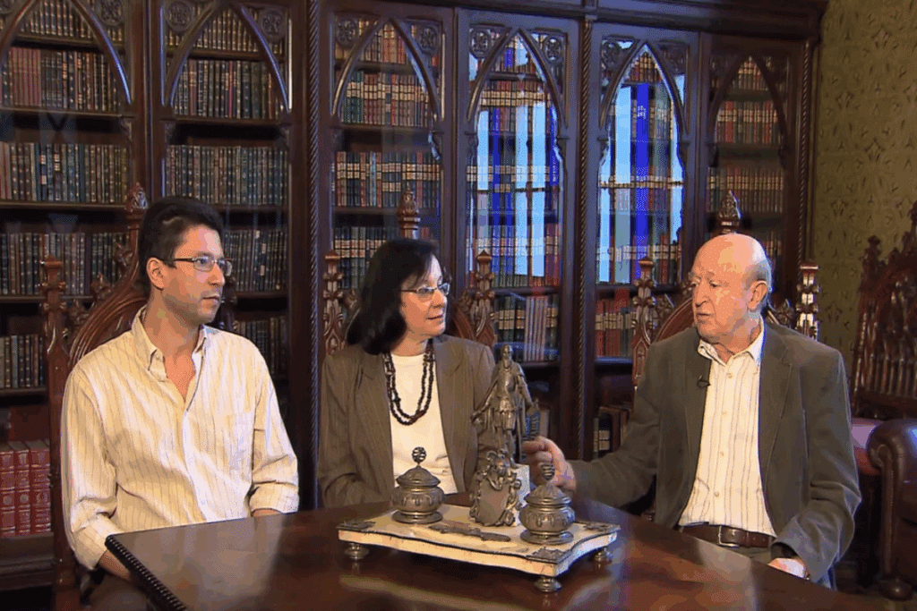 Uma cena de entrevista mostra três pessoas sentadas ao redor de uma mesa de madeira em uma biblioteca. Ao fundo, há grandes estantes repletas de livros, com detalhes de madeira escura e janelas de vidro. Sobre a mesa, há objetos decorativos antigos. Os três conversam em um ambiente com aparência clássica e formal.