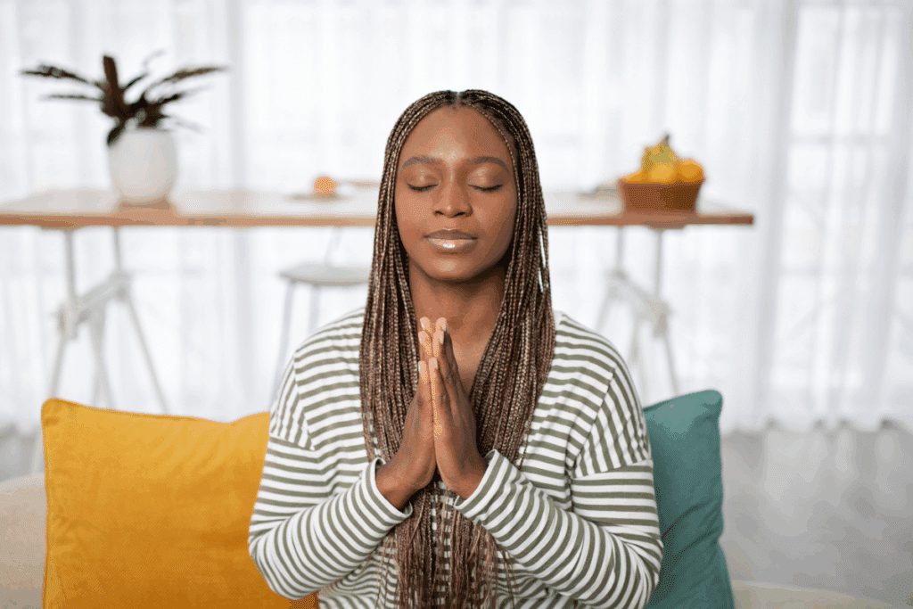 Uma mulher negra com longas tranças (box braids) está sentada em um sofá, de olhos fechados e mãos postas em posição de oração ou meditação. Ela veste uma blusa de manga longa listrada em verde e branco
