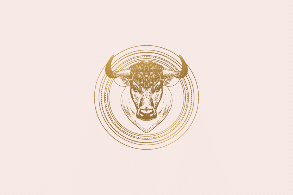 Ilustração do signo Touro: gravura dourada da cabeça de um touro vista de frente, com chifres curvados para cima que saem do medalhão de anéis dourados finos e pontilhados; em fundo rosa-claro.