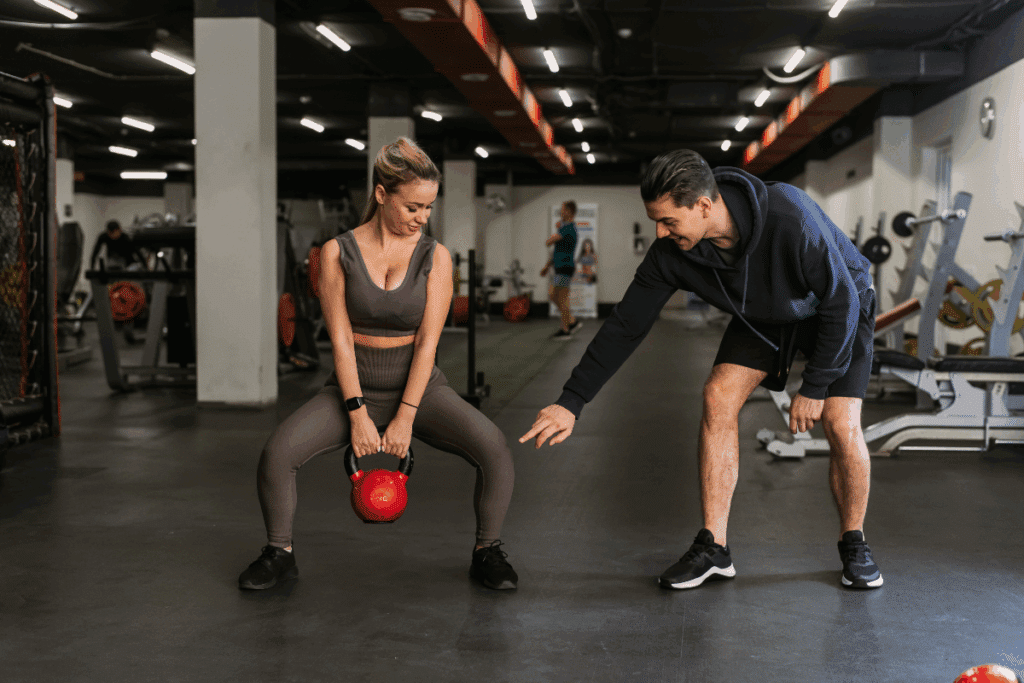 Mulher de cabelos loiros presos em um rabo de cavalo, vestindo top esportivo cinza-escuro, legging da mesma cor e tênis pretos, realiza um agachamento segurando um peso vermelho entre as pernas. Ao lado, um homem de cabelos pretos penteados para trás, usando moletom azul-marinho, shorts pretos e tênis escuros, inclina o corpo e aponta para o peso enquanto acompanha o exercício. Os dois estão em uma academia com piso escuro, equipamentos de musculação, anilhas e aparelhos ao fundo. O teto é preto, com luminárias lineares que iluminam o ambiente.