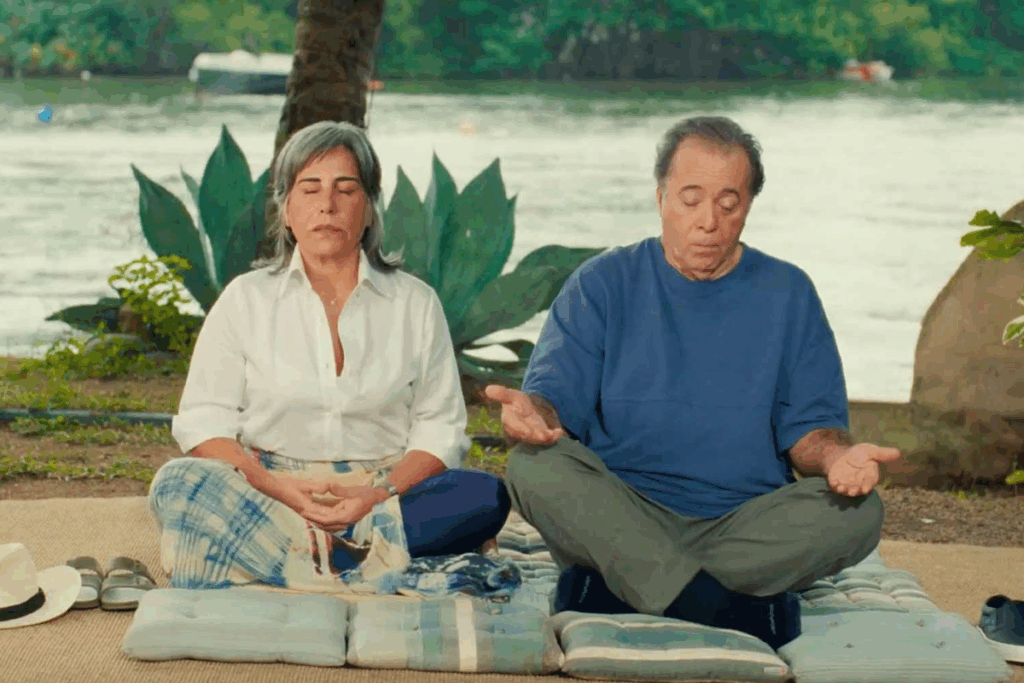 Cena de filme em que uma mulher de cabelos grisalhos e camisa branca e um homem de camiseta azul meditam de olhos fechados, sentados em almofadas sobre um tapete. Ao lado deles há um chapéu panamá e dois pares de sapatos. Ao fundo aparecem um rio, plantas e barcos.