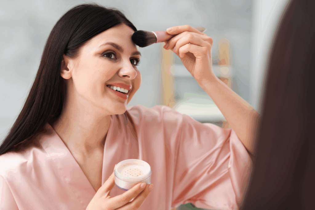 Mulher de cabelos escuros, longos e lisos, sorrindo diante do espelho enquanto passa pó na testa com um pincel de cerdas escuras e cabo rosa. Na outra mão, ela segura um pote de pó solto claro. Ela veste um robe de cetim rosa-claro, e o fundo é um banheiro claro desfocado.